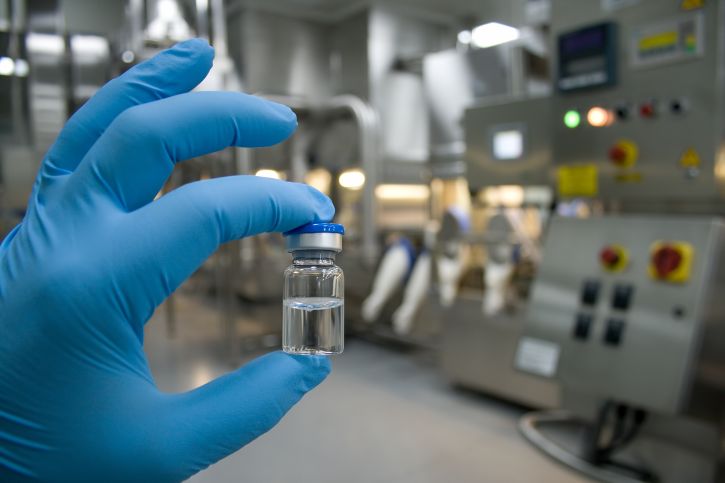 Hand in Handschuh die Ampulle mit Flüssigkeit hält, im Hintergrund Pharma-Anlage; Biopharmazeutika, Biopharma, Biotechnologie, Biologicals