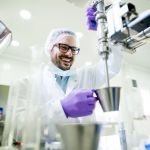 Young smiling researcher with gloves and hair protection working on the lab machine. Der jährlichen Umfrage des Biotechverbands BIO Deutschland zufolge, bewerten Unternehmen der Branche ihre aktuelle und zukünftige Situation besser als im Vorjahr. Die Indices sind jedoch deutlich niedriger als in der Hochphase der Corona-Pandemie.